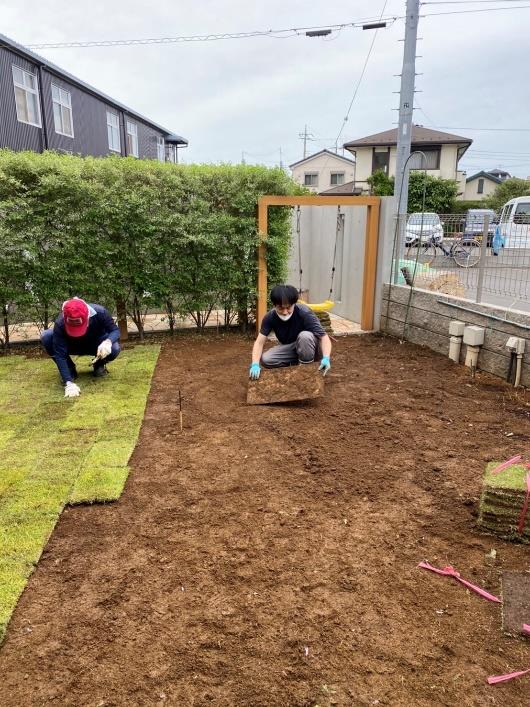 改良した土の上にしゃがんで芝生を並べる作業員2人の写真（右が茄子男）