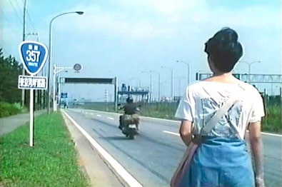 道路脇に立つ人物とバイクが通り過ぎる様子のレトロな写真